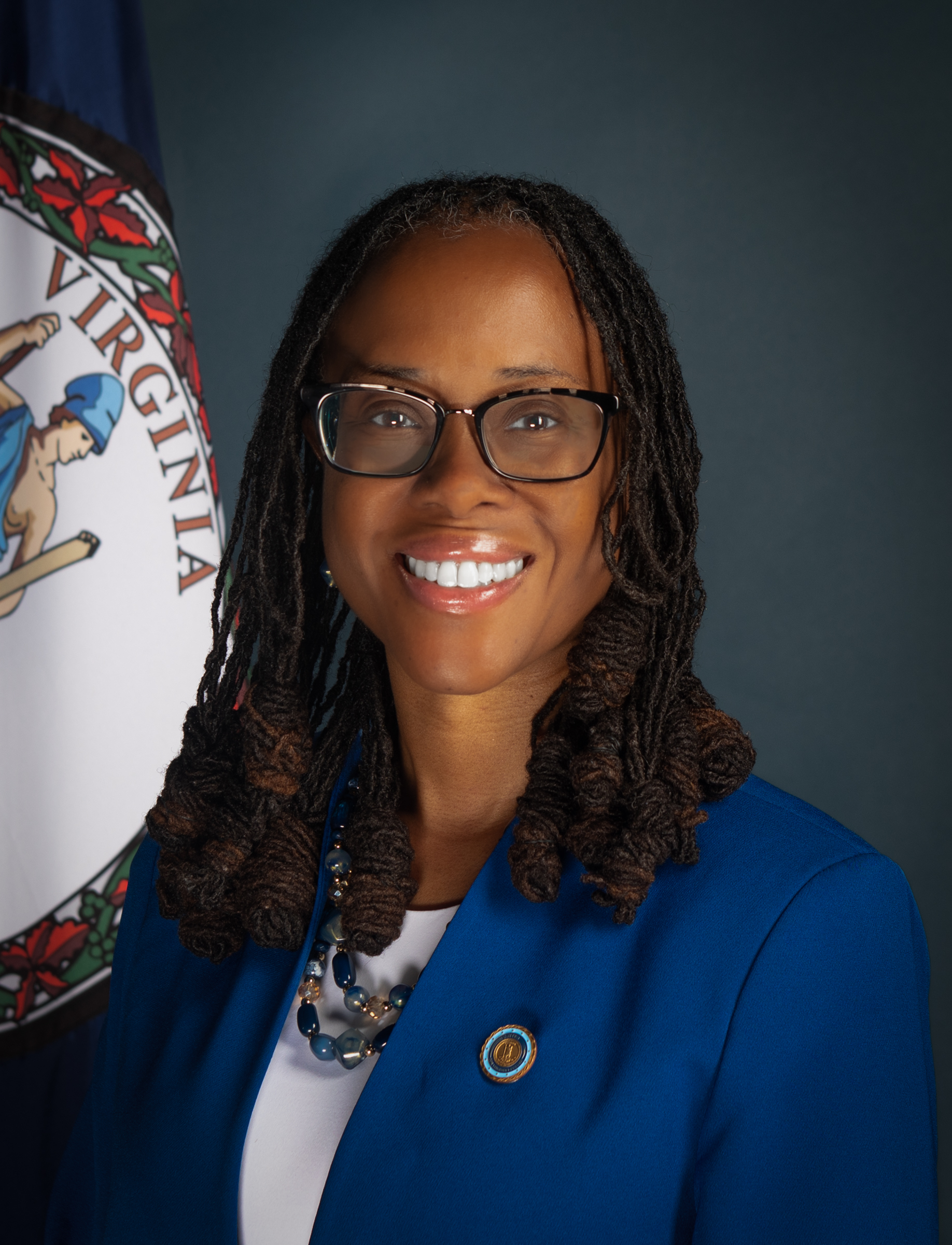 A woman with glasses and dark brown hair in front of the virginia flag