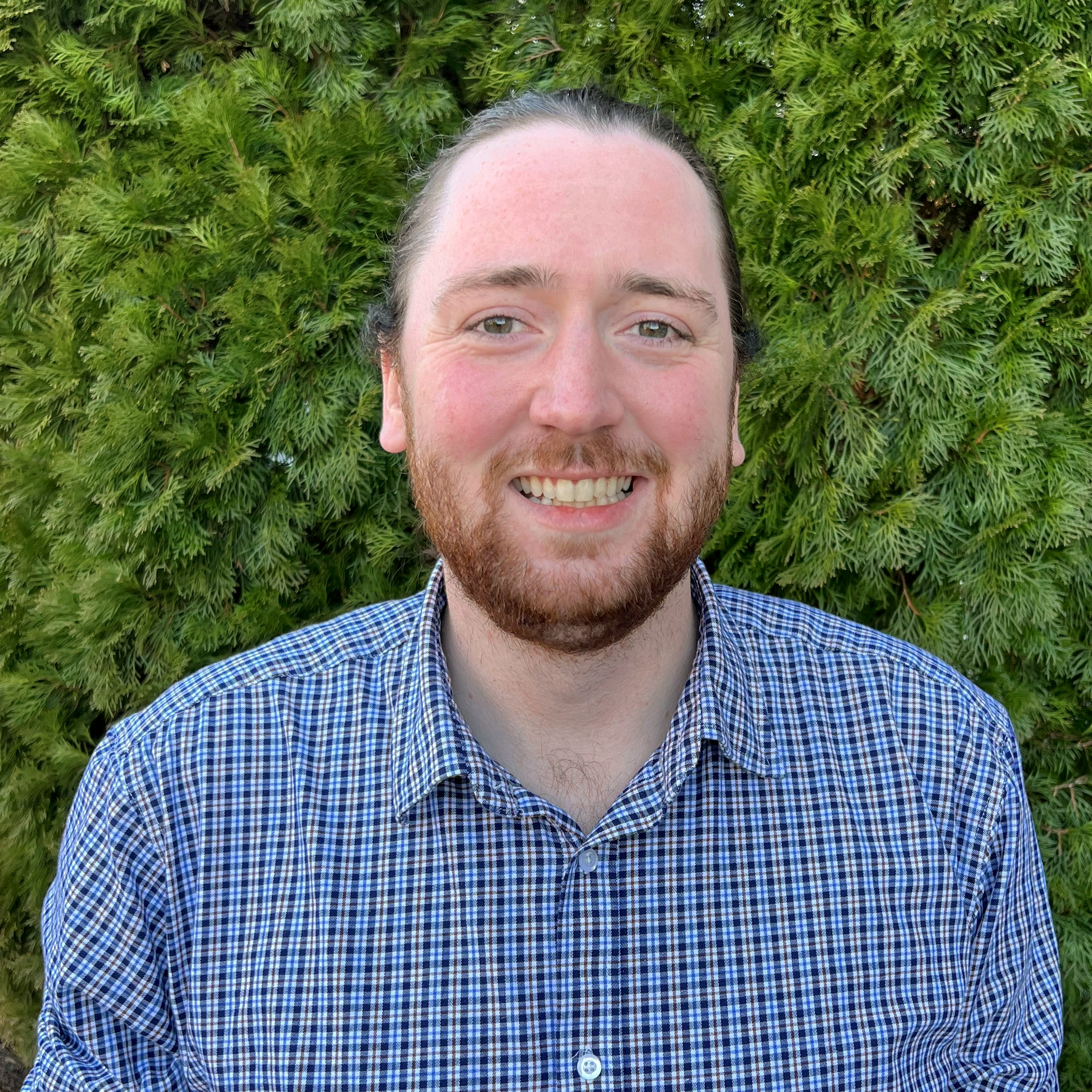 Ben Cook, senior software engineer, wears a blue button up in his headshot and stands in front of a bush